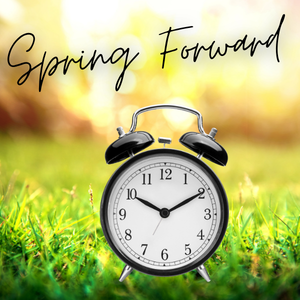 Black vintage alarm clock sitting in green grass with soft golden sunlight in the background, with "Spring Forward" in black script lettering above it.