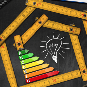 A folding ruler arranged in the shape of a house surrounds an energy efficiency rating scale and a lightbulb illustration, with a calculator and tools on a dark background.